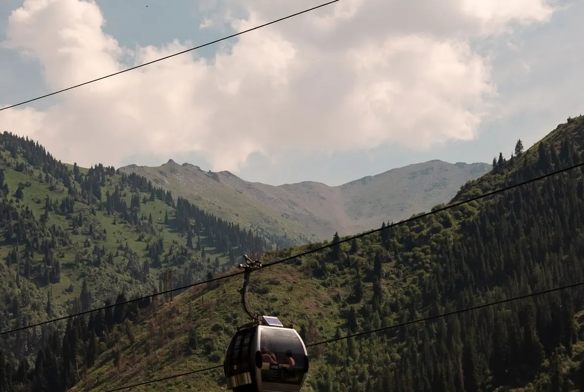 A cable-car gondola gliding above a green, spruce-covered mountain gorge under summer clouds